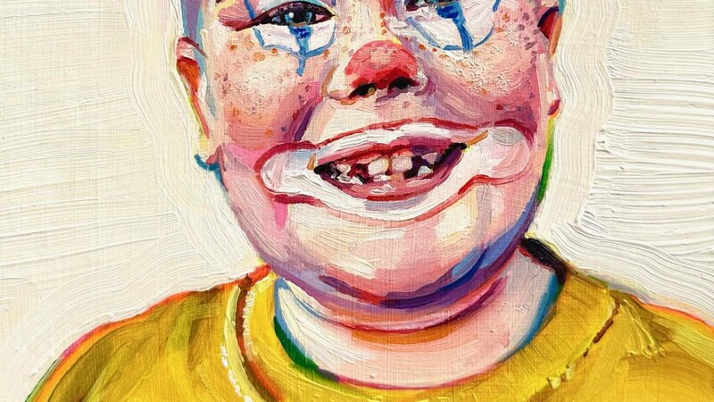A young boy in a yellow shirt with clown makeup. He's bald and is missing teeth. The paint is chunky and colorful. 