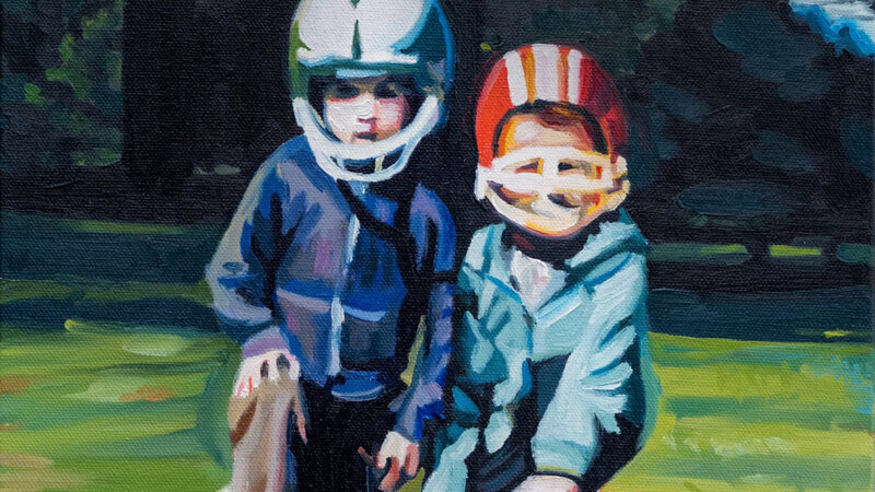 Two brothers kneeling down posting with their football helmets and a football. They look serious, but their helmets are too big. There is greenery in the background. 