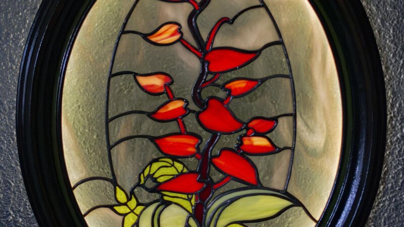 An image of a stained glass panel illuminated from behind. It shows a red-leafed plant in a smoky grey background with green foliage and a small snail in the foreground.