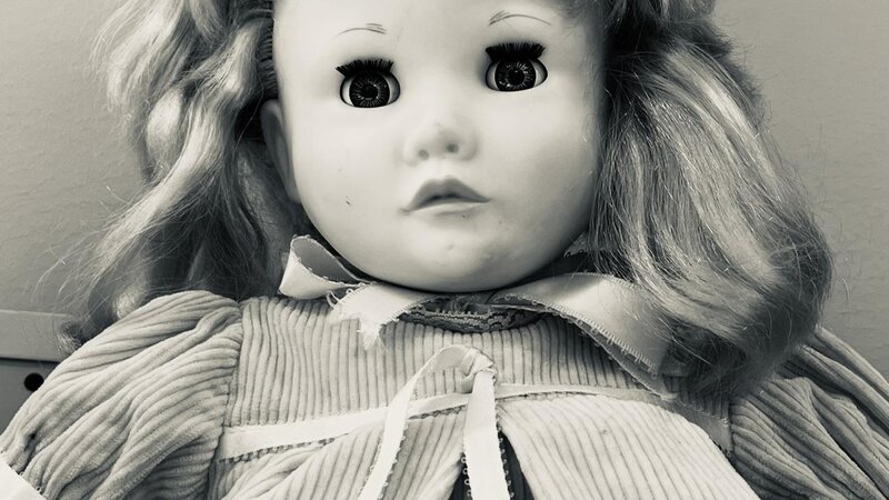 A child's baby-doll staring creepily from the shelf of a second hand store. This image is in black and white.