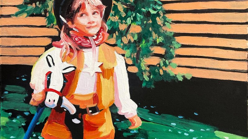 A little girl dressed like a cowgirl, looking up to the left. The background is a simple mix of greenery and siding on a house. 