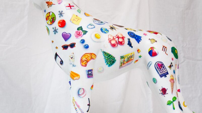 A white dog sculpture painted with dozens of tiny little objects, such as hearts, fruit, plants, logos, toys, etc. 