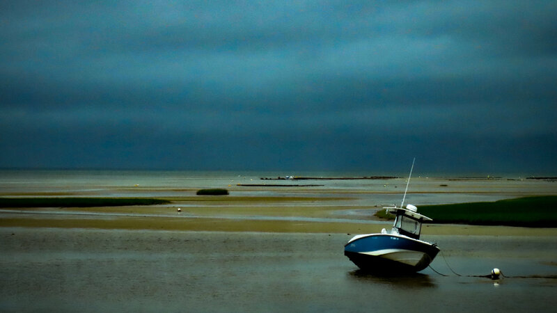 Photograph of a Stormy day on a cape cod Beach. The sky is dark gray and blue. A boat is tethered to an off shore mourning that is almost out of the water because the tide is so low. 
