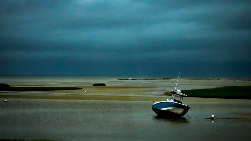 Photograph of a Stormy day on a cape cod Beach. The sky is dark gray and blue. A boat is tethered to an off shore mourning that is almost out of the water because the tide is so low. 