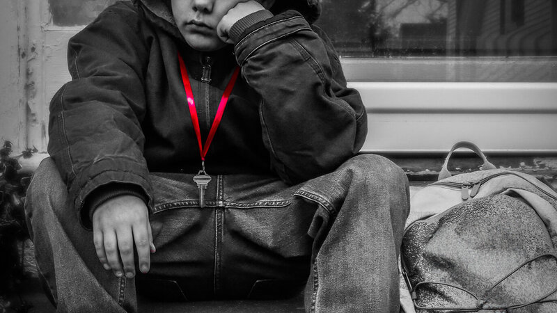 Little boy who appears sad sits on stoop with red ribbon holding key around his neck 