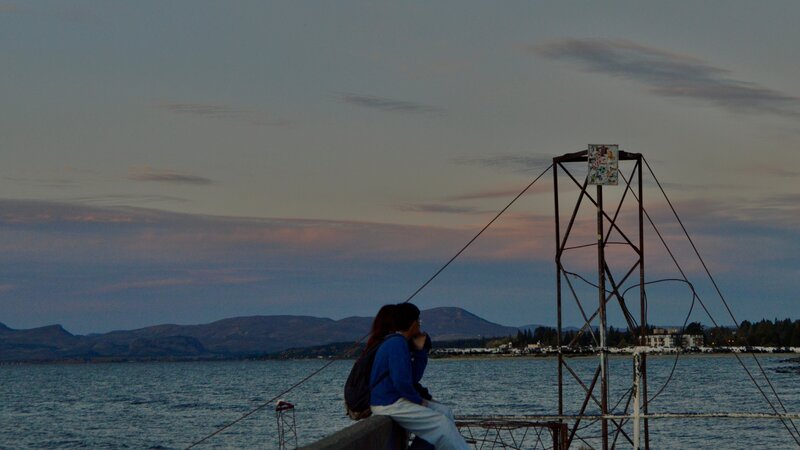 Image of two people sat beneath the sunset, under the moon, with mountains in the distance at the end of a pier in Bariloche, Argentina.