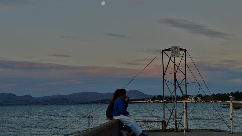 Image of two people sat beneath the sunset, under the moon, with mountains in the distance at the end of a pier in Bariloche, Argentina.