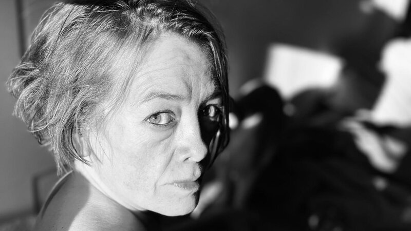 Photograph of a person looking over their shoulder as they sit in the sun. The image is black and white the shadows in the background draw your attention to the persons face. The person has short messy hair and is looking directly at the viewer.