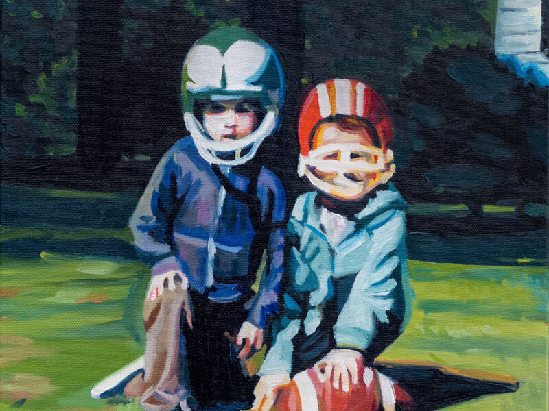 Two brothers kneeling down posting with their football helmets and a football. They look serious, but their helmets are too big. There is greenery in the background. 