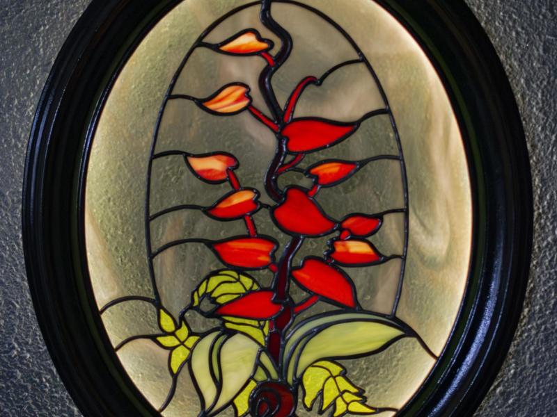 An image of a stained glass panel illuminated from behind. It shows a red-leafed plant in a smoky grey background with green foliage and a small snail in the foreground.