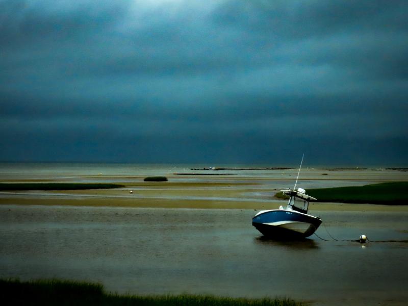 Photograph of a Stormy day on a cape cod Beach. The sky is dark gray and blue. A boat is tethered to an off shore mourning that is almost out of the water because the tide is so low. 