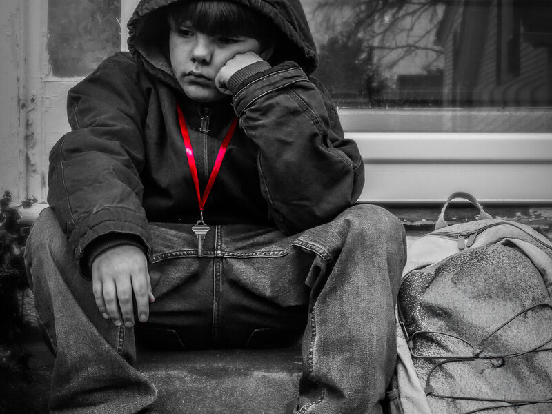 Little boy who appears sad sits on stoop with red ribbon holding key around his neck 