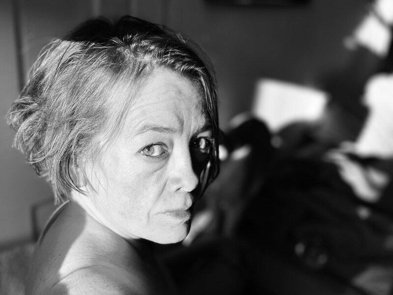 Photograph of a person looking over their shoulder as they sit in the sun. The image is black and white the shadows in the background draw your attention to the persons face. The person has short messy hair and is looking directly at the viewer.