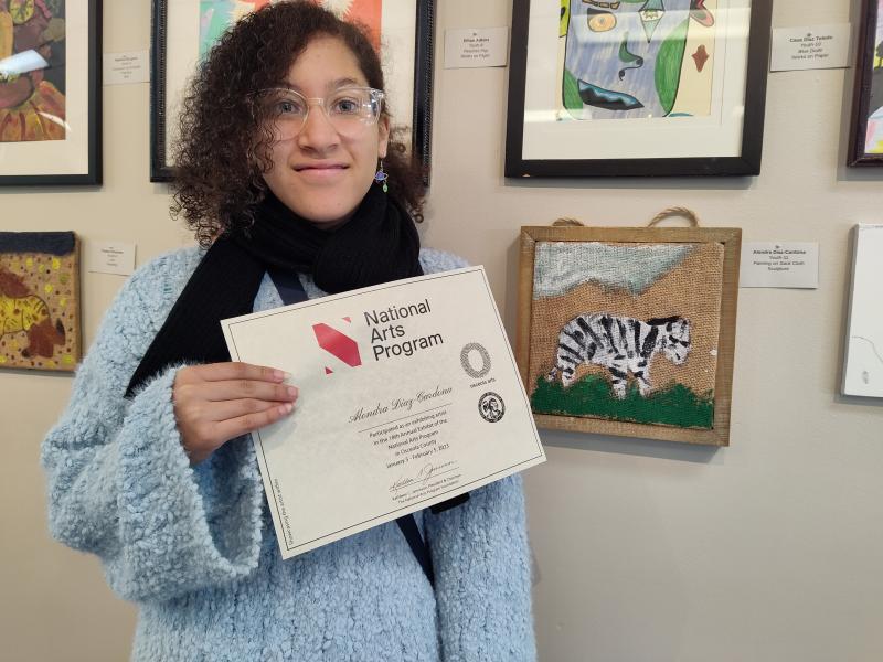 artist participant holding her certificate in front of her art on the wall