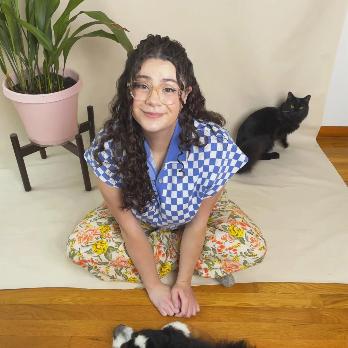 Artist Michelle Hawran sitting on the ground with her black cat smiling up at the camera. There is a plant behind her. 