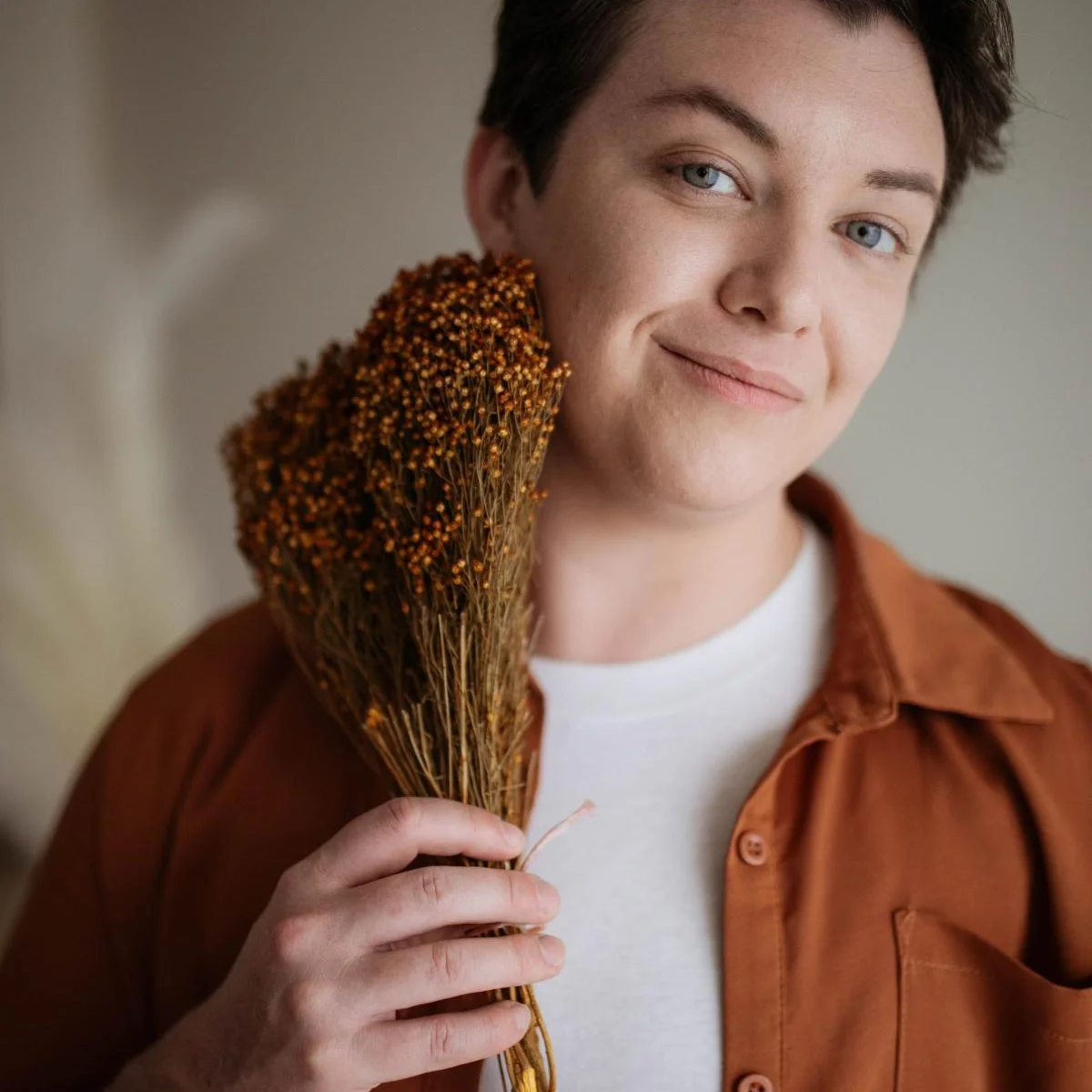 Milo, a white person with short dark hair and blue eyes, is pictured in an auburn shirt over a white tee. They are holding a dried bouquet of flowers and smiling