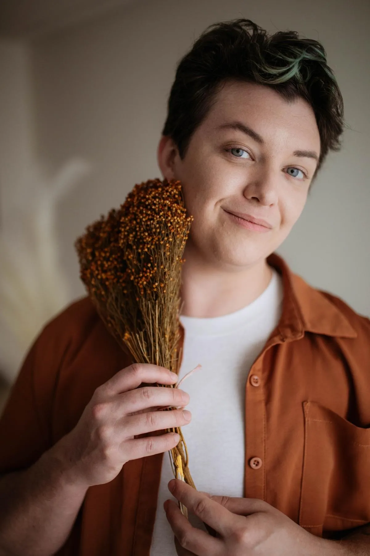 Milo, a white person with short dark hair and blue eyes, is pictured in an auburn shirt over a white tee. They are holding a dried bouquet of flowers and smiling