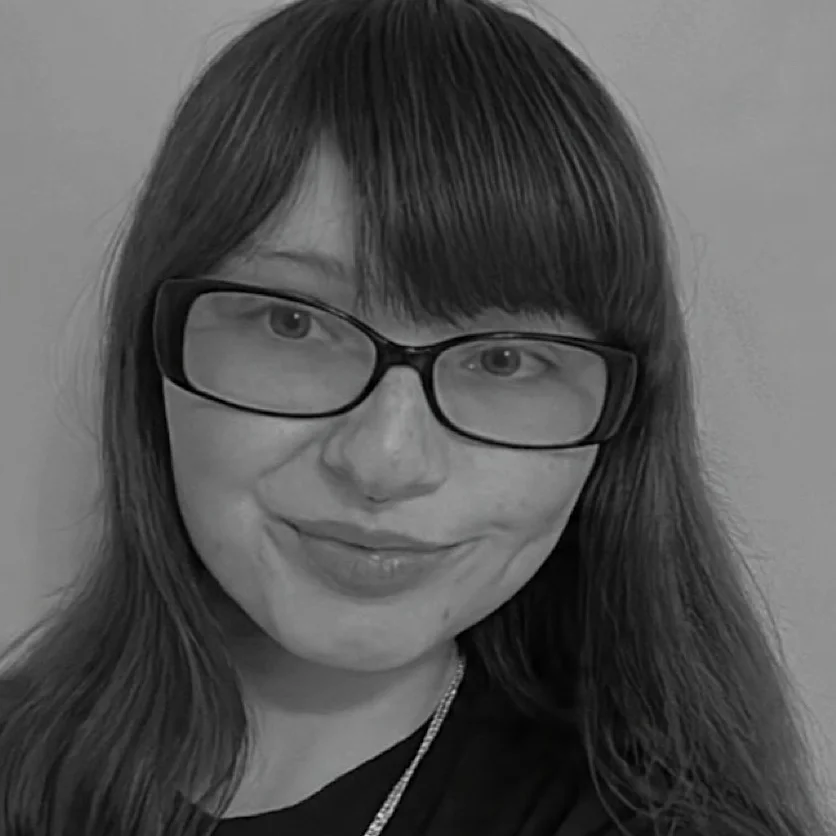 The artist has long hair with bangs, short rectangular glasses, and a necklace 