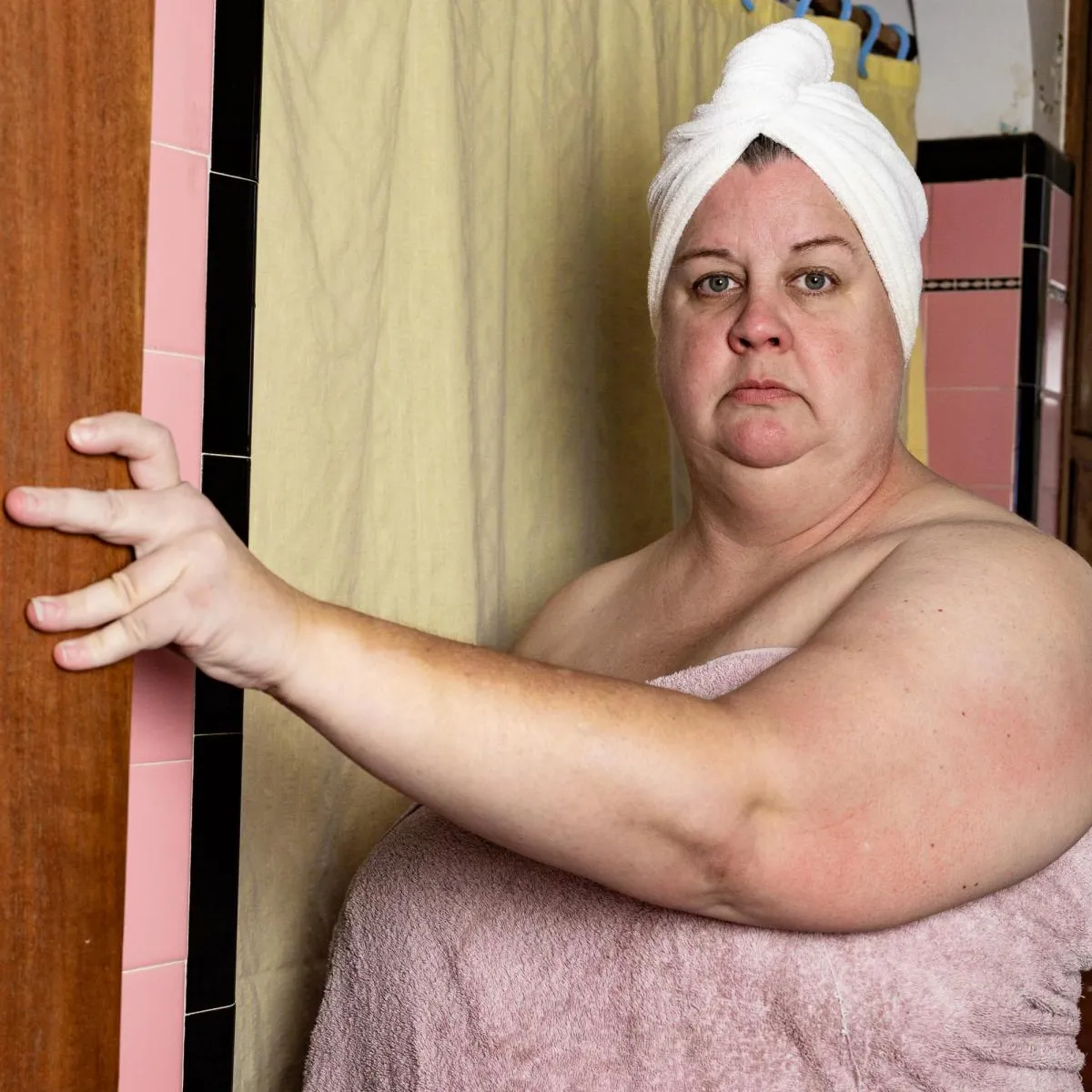 Middle-aged woman staring directly at the camera wearing a pink bath sheet and white hair wrap standing outside of the shower with a yellow shower curtain in a pink and black tiled bathroom.