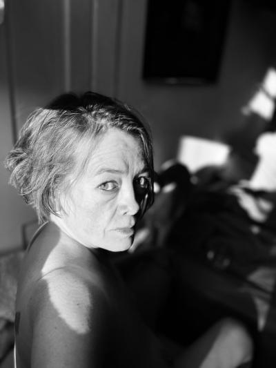 Photograph of a person looking over their shoulder as they sit in the sun. The image is black and white the shadows in the background draw your attention to the persons face. The person has short messy hair and is looking directly at the viewer.
