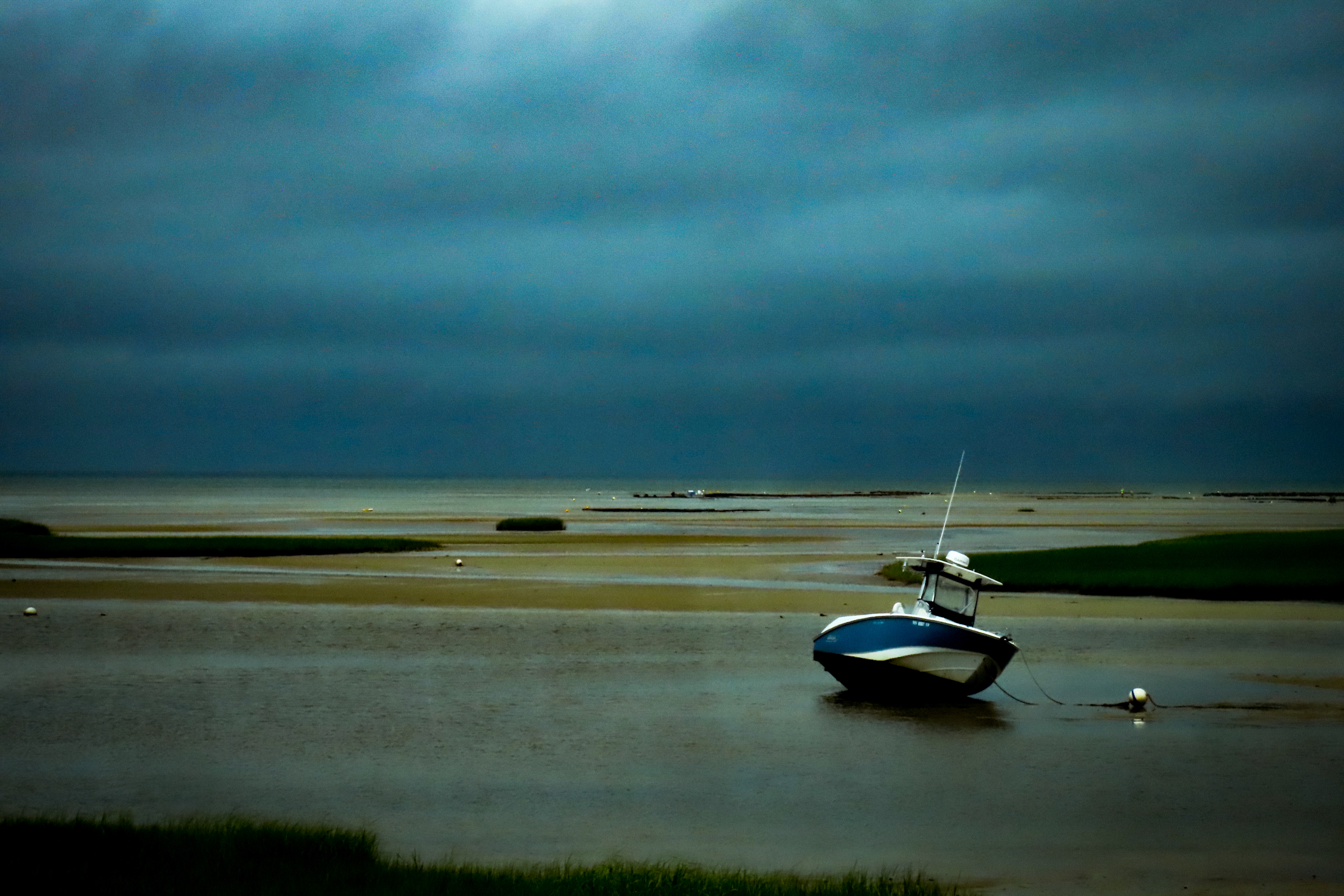 Photograph of a Stormy day on a cape cod Beach. The sky is dark gray and blue. A boat is tethered to an off shore mourning that is almost out of the water because the tide is so low. 