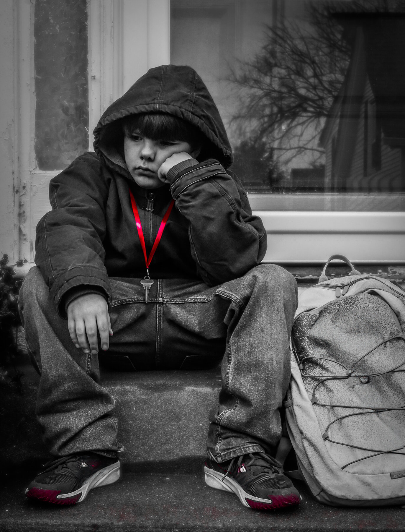 Little boy who appears sad sits on stoop with red ribbon holding key around his neck 