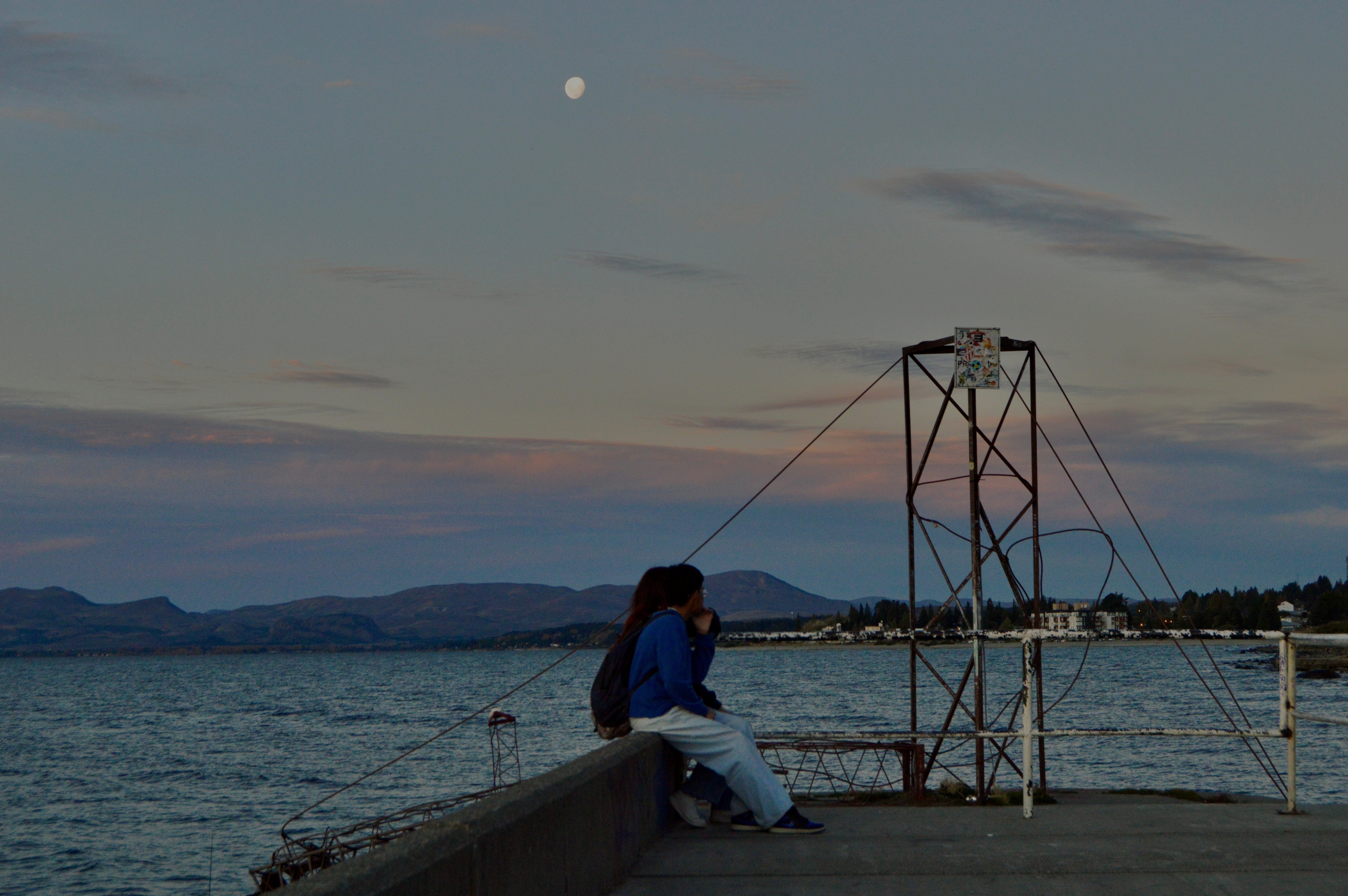 Image of two people sat beneath the sunset, under the moon, with mountains in the distance at the end of a pier in Bariloche, Argentina.