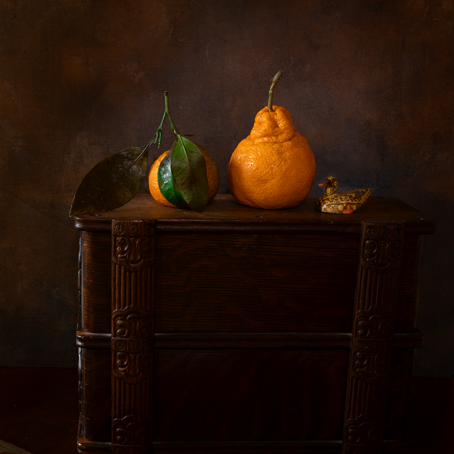 Still life image showing 2 mandarin oranges and a papier mache duck on top of a piece of furniture.